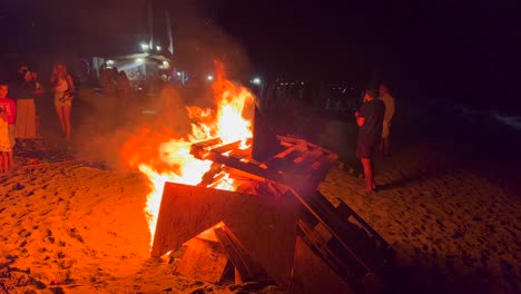 Traditional-bonfire-summer-festival-at-the-beach-at-the-San-Juan-celebration-in-Marbella-Spain,-friends-and-family-enjoying-a-fun-party,-big-burning-fire-and-hot-flames-at-night,-4K-shot