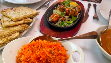 Traditional-Indian-Tandoori-chicken-with-onions-on-a-very-hot-plate,-butter-chicken,-cheese-naan-bread-and-coconut-rice-in-a-restaurant,-authentic-asian-food,-4K-shot