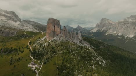 Aerial-view-aproaching-massive-rock-formations-with-distant-tall-mountains-in-the-background,-green-fields-at-the-bottom,-cloudy-day,-cinematic-color-grade
