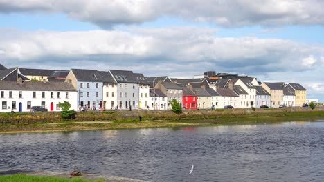 Pretty-little-houses-of-different-colours-along-a-river-edge-with-birds-flying,-Galway