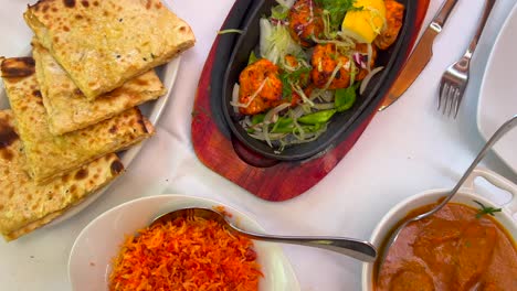 Traditional-Indian-Tandoori-chicken-with-onions-on-a-very-hot-plate,-butter-chicken,-cheese-naan-bread-and-coconut-rice-in-a-restaurant,-authentic-asian-food,-4K-top-shot