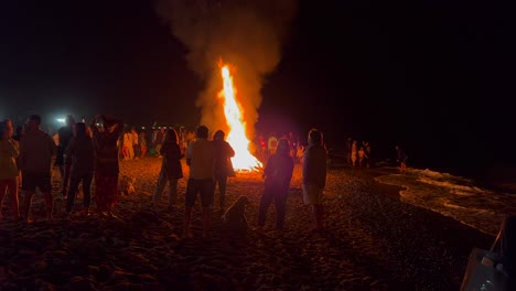 People-standing-around-a-traditional-bonfire-summer-festival-at-the-beach-at-the-San-Juan-celebration-in-Marbella-Spain,-enjoying-a-fun-party,-big-burning-fire-and-hot-flames-at-night,-4K-shot