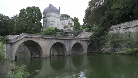 Green-water-and-trees-in-surrounding-park-in-Chateauneauf-sur-Loire,-with-stone-arched-bridge-on-an-overcast-day