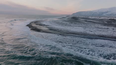 Aerial-landscape-view-of-ocean-waves-crashing-on-to-diamond-beach,-covered-in-snow,-at-sunset