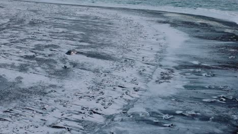 Aerial-view-over-ocean-waves-crashing-on-to-diamond-beach,-covered-in-snow,-at-sunset