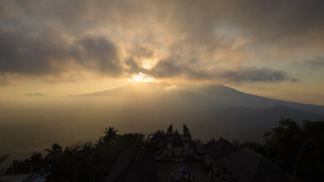 Mount-Agung-volcano-during-golden-hour-sunset-sun-beams-with-dramatic-cloud-motion,-gate-of-heaven-silhouetted-in-foreground