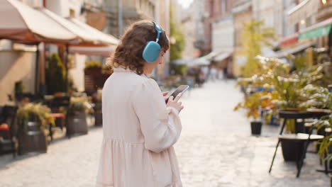 Happy-young-woman-in-wireless-headphones-choosing,-listening-music-in-smartphone-dancing-outdoors