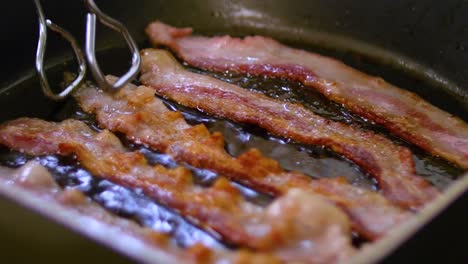 Bacon-strips-sizzling-in-frying-are-flipped-by-tongs,-closeup