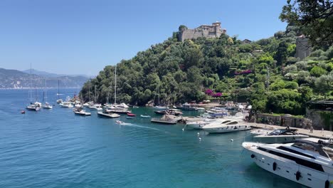 Panning-shot-of-scenic-views-of-colourful-houses,-Castello-Brown,-a-historic-castle-perched-on-a-hill-and-boats-sailing-and-mooring-in-the-small-harbour-of-Portofino,-Italy