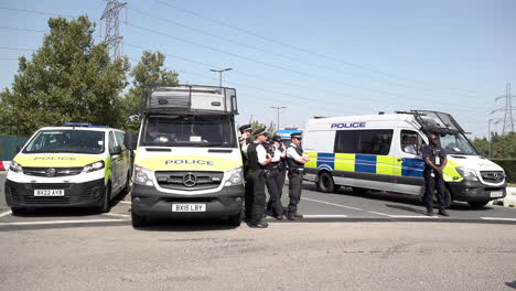 A-unit-of-Metropolitan-officers-stand-by-three-police-vehicles-that-are-blocking-a-road-on-a-bright-and-hot-sunny-day