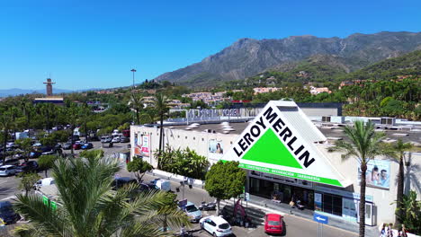 Aerial-view-of-Leroy-Merlin-home-improvement-and-gardening-retailer,-big-hardware-store-parking-with-palm-trees-and-mountain-view-in-Marbella-Malaga-Spain,-4K-shot