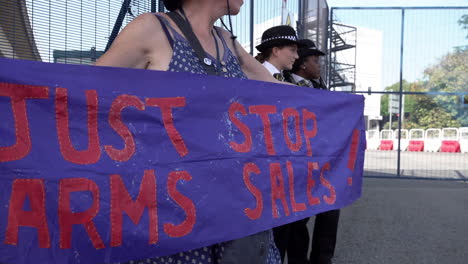 A-protestor-stands-next-to-two-police-officers-while-holding-a-purple-banner-with-red-writing-that-reads,-“Just-stop-arms-sales