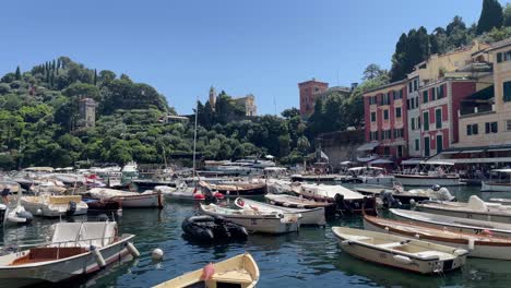 Boats-mooring-side-by-side-in-the-small-harbour-of-Portofino,-Italy-and-the-background-view-of-the-iconic-landmarks-of-Chiesa-di-San-Giorgio