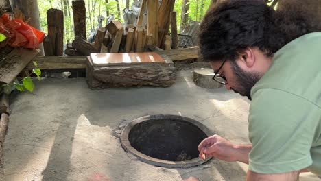 a-man-is-trying-to-light-matches-to-fire-the-traditional-oven-clay-oven-round-circle-shape-in-ground-works-with-wood-fire-charcoal-bone-fire-camp-in-order-to-bake-delicious-original-Turkish-flat-bread