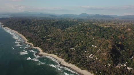 High-altitude-aerial-footage-of-a-tropical-beach,-during-daylight,-with-tropical-forest-and-mountains-in-the-background