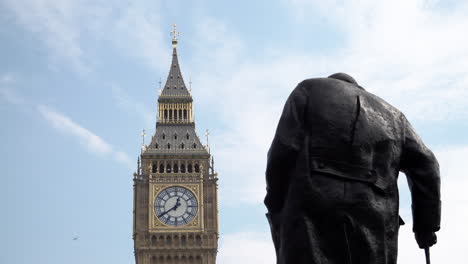 A-private-jet-plane-flies-behind-the-Winston-Churchill-statue-on-Parliament-Square-and-the-Elizabeth-Tower-at-the-Palace-of-Westminster,-that-houses-the-Big-Ben-bell-on-a-bright-and-hot-day