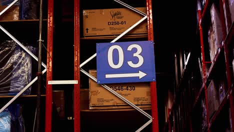 A-Large-03-Warehouse-Logistics-Distribution-Center-Sign-with-an-Arrow-Points-Right-Down-a-Set-of-Factory-Storage-Racks-Containing-Ashley-Signature-Furniture-Boxes-of-Merchandise-and-Products
