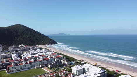 Aerial-view-of-Praia-Brava,-Florianópolis,-featuring-beachfront-buildings,-pristine-shoreline,-majestic-mountains,-and-a-clear-blue-sky