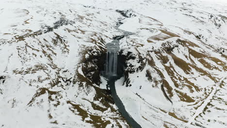 Drone-shot-circling-the-wintry-Skogafoss-waterfall,-snowy,-overcast-day-in-Iceland