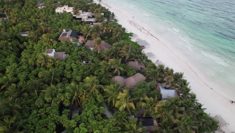 Aerial-orbit-top-down-shot-of-cabins-and-huts-surrounded-by-palm-trees-in-a-white-sand-beach-with-crystal-clear-blue-water-in-Tulum,-Mexico