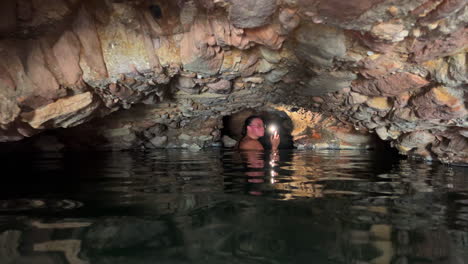 Girl-exploring-with-flashlight-dark-caves-of-Roman-Baths-of-Casares-Manilva,-people-swimming-in-natural-spring-healing-sulfur-water-in-Spain,-good-health-benefits,-4K-shot