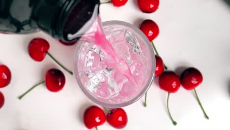 Close-up-birdseye-shot-of-a-shaker-pouring-a-tasty-cherry-whey-to-clear-ice-cubes-while-cherries-lie-on-the-side