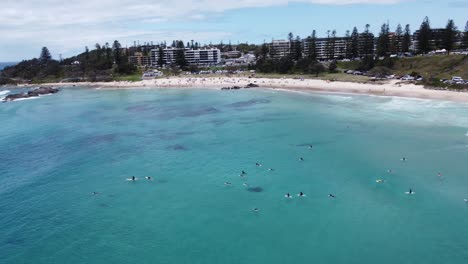 Drone-Volando-Sobre-Surfistas-Mostrando-Una-Hermosa-Playa-En-Australia-Y-Una-Ciudad-Costera-Al-Fondo