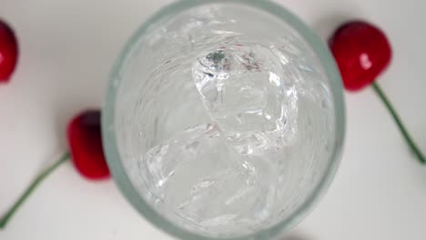 Birdseye-close-up-shot-of-clear-ice-cubes-falling-into-the-glass-for-a-cool-drink-while-cherries-lie-on-the-side-in-slow-motion