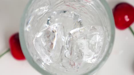 Close-up-birdseye-shot-of-clear-ice-cubes-falling-into-a-beautiful-glass-for-a-cool-drink-while-cherries-lie-on-the-side-in-slow-motion