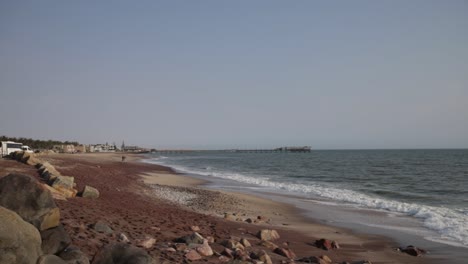 Ein-Strand-Mit-Rotem,-Schwarzem-Und-Weißem-Sand-Und-Verschiedenen-Felsenarten-Mit-Dem-Swakopmund-Steg-Im-Hintergrund