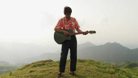 Asian-male-musician-enjoying-strumming-guitar-and-singing-on-mountain-top