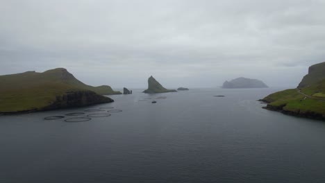 Drone-flying-towards-Drangarnir-and-Tindholmur-through-a-salmon-farm-in-Faroe-Islands