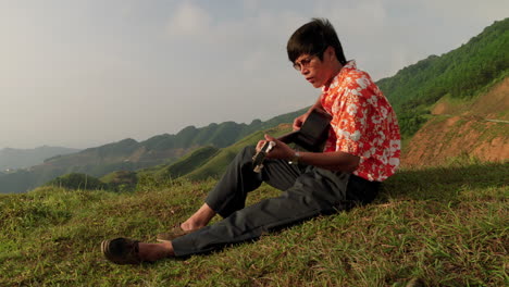 Asian-male-musician-busy-composing-song-and-strumming-guitar-on-hill-top