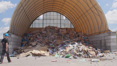 Worker-walks-toward-pile-of-garbage-at-recycling-facility,-wide-slomo