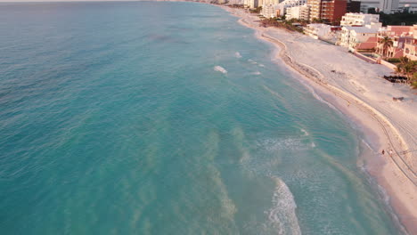 Aerial-shot-of-rolling-waves-in-a-white-sand-beach-surrounded-by-resorts-and-hotels-in-a-crystal-clear-blue-ocean-in-Cancun,-Mexico