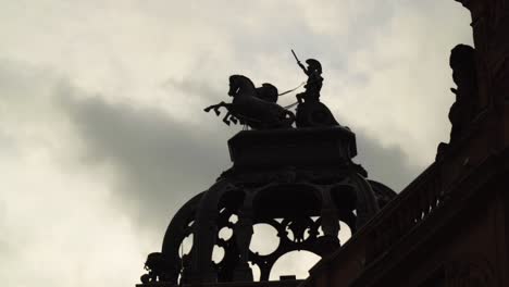 powerful-cinematic-scene-of-a-roman-warrior-riding-a-chariot-with-two-horses-raised-arm-holding-a-sword-in-the-sky-sculpture-on-top-of-a-roof-silhouette-dramatic-clouds-moving-quick-speed-background