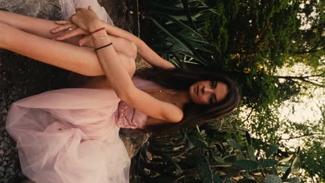 Horizontal-shot-of-full-body-portrait-of-gorgeous-young-brunette-woman-sitting-on-ground-wearing-pink-gown-in-a-garden