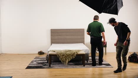 Two-People-Staging-a-Furniture-a-Bed-Frame-Backstage-Inside-a-Professional-Hard-Wood-Photography-Film-Set-Studio-Surrounded-by-Bright-Lights-Softbox-for-a-Commercial-Video-Production