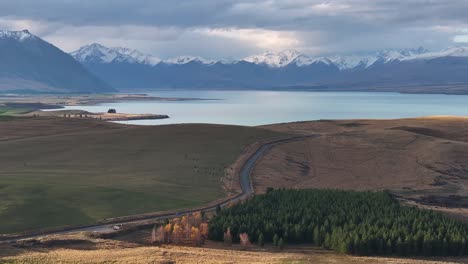 Aerial-view-of-road-alongside-Lake-Tekapo-and-snowy-peaks-of-Southern-Alps,-New-Zealand-landscape