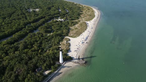 Cape-Florida-lighthouse-and-beach,-aerial-descend-dolly-tilt-up-to-golden-sandy-stretch-and-city-skyline