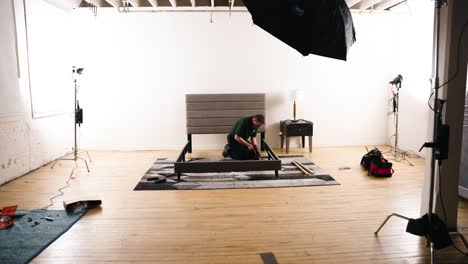 Camera-Dolly-in-Professional-Carpentry-Man-Assembling-Furniture-a-Bed-Frame-Backstage-Inside-a-Professional-Hard-Wood-Photography-Film-Set-Studio-Surrounded-by-Bright-Lights-Softbox