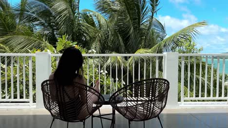 Rückansicht-Eines-Mädchens-Im-Schwarzen-Badeanzug,-Das-An-Einem-Sonnigen-Tag-Auf-Einem-Luxuriösen-Balkon-Sitzt-Und-Den-Blick-Auf-Die-Palmen-Bewundert