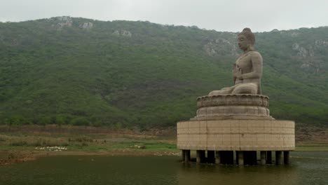 70-foot-high-Buddha-statue-at-Ghora-Katora-lake-surrounded-by-mountains-under-hazy-sky,-Rajgir,-Bihar,-India