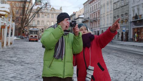 Senior-wife-husband-tourists-taking-photo-pictures-on-retro-camera,-walking-on-winter-city-street