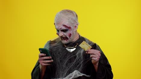 Sinister-man-Halloween-zombie-using-credit-bank-card-and-smartphone-while-purchases-online-shopping