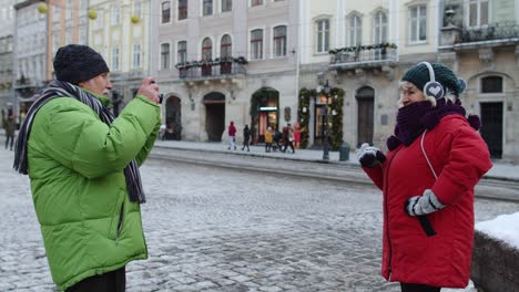 Senior-wife-posing-for-camera-laughing-while-husband-is-taking-photo-pictures-on-winter-city-street