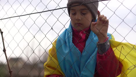 sad-little-girl-with-the-flag-of-Ukraine-behind-a-metal-fence.-Social-problem-of-refugees-and-forced-migrants