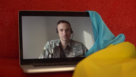 Soldier-in-military-uniform-sits-on-sofa-at-home-using-webcam-laptop-for-communication-with-family.-Ukrainian-flag-on-the-background.-Military-actions-in-Ukraine.-Free-democratic-people-in-the-state
