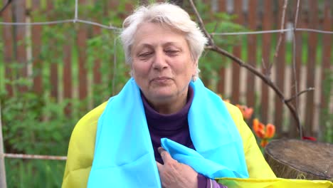 Senior-white-haired-woman-wearing-eyeglasses-standing-outdoors-in-the-country-waving-the-Ukrainian-flag-looking-away-feeling-the-freedom.-No-war,-stop-fights,-we-want-peace.