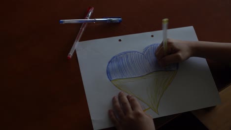 Die-Kinderhände-Eines-Kindes-Zeichnen-Ein-Herzförmiges-Symbol-Mit-Dem-Bild-Der-Nationalflagge-Der-Ukraine.-Sicht-Von-Oben.-Kinder-Gegen-Den-Krieg.-Kinderzeichnung-Für-Den-Frieden-In-Der-Ukraine